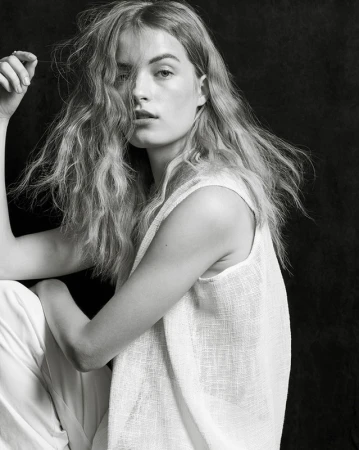 Portrait of a woman with tousled long hair wearing a white sleeveless top against a dark textured background.