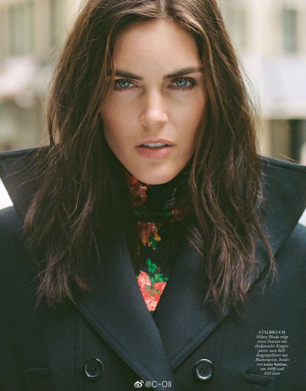 Portrait of an adult woman with long brunette hair, wearing a black wool overcoat and floral top, looking directly at the camera outdoors.