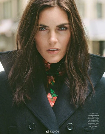 Portrait of an adult woman with long brunette hair, wearing a black wool overcoat and floral top, looking directly at the camera outdoors.