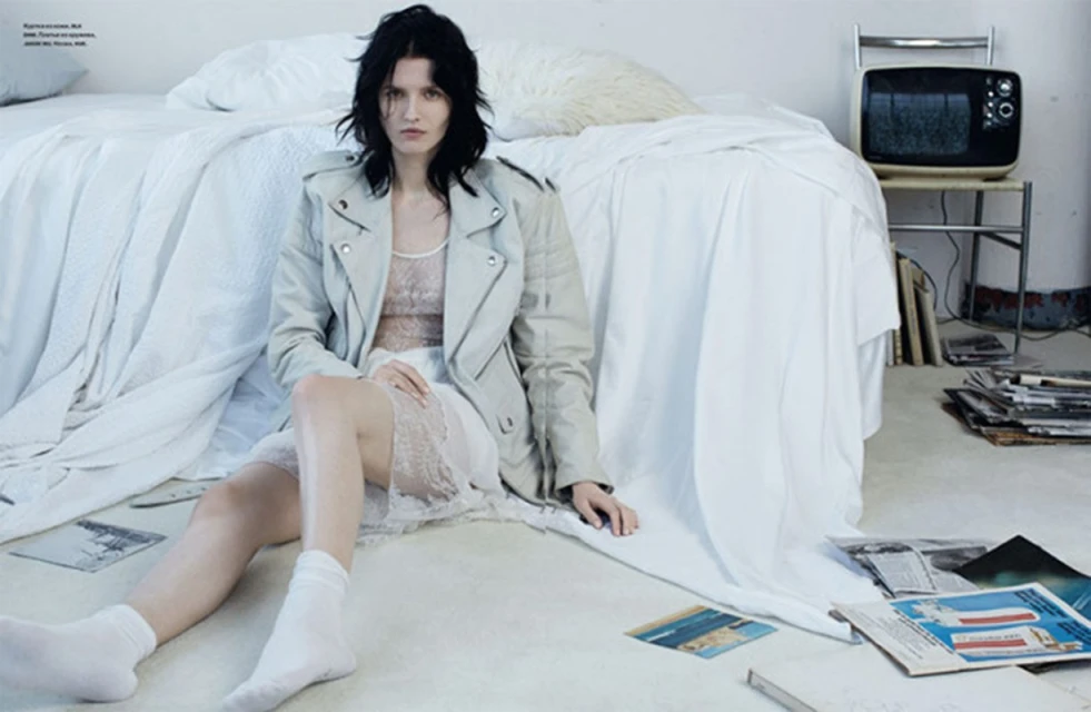 A woman with dark hair sits on the floor wearing a jacket and dress next to a bed and retro television in an indoor setting.