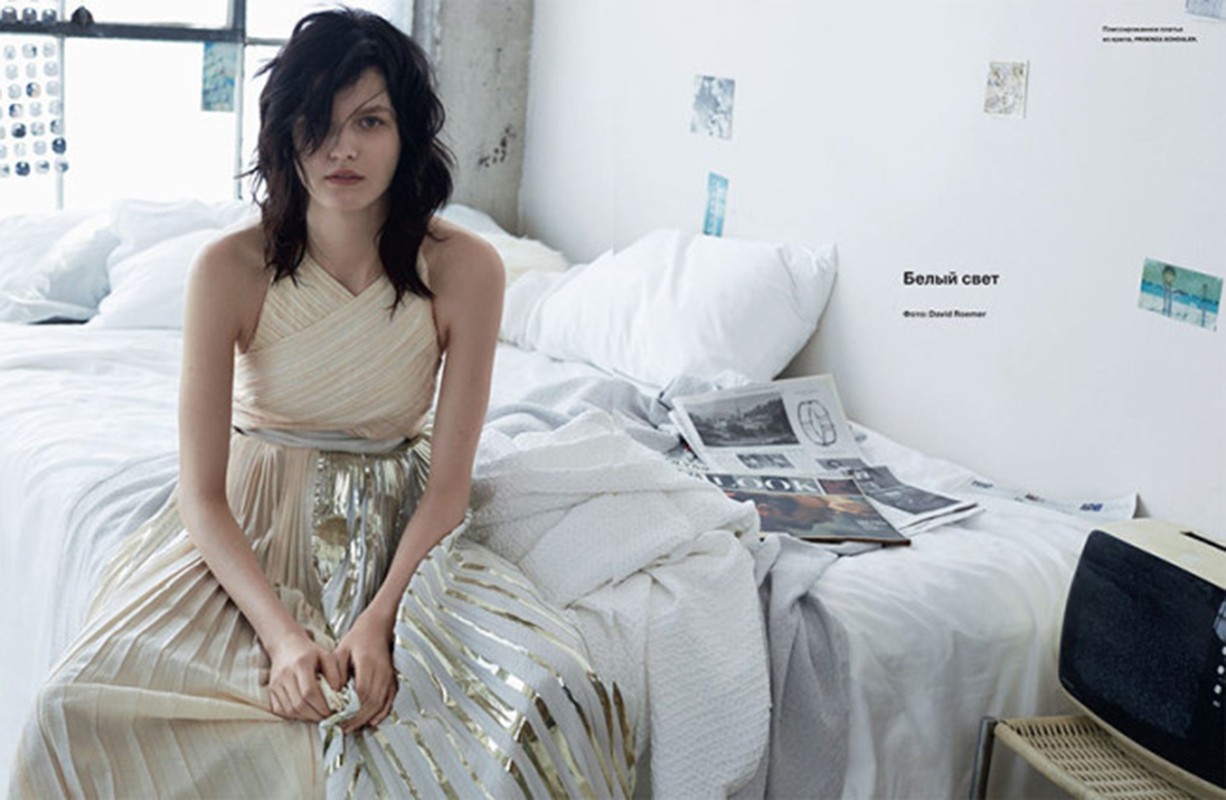 A woman with dark hair sits on a bed in an off-white and gold dress. Natural light fills the room, highlighting the soft textures.