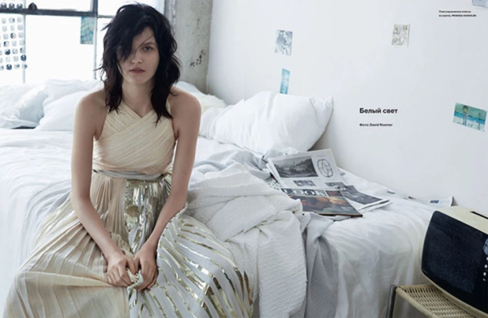 A woman with dark hair sits on a bed in an off-white and gold dress. Natural light fills the room, highlighting the soft textures.