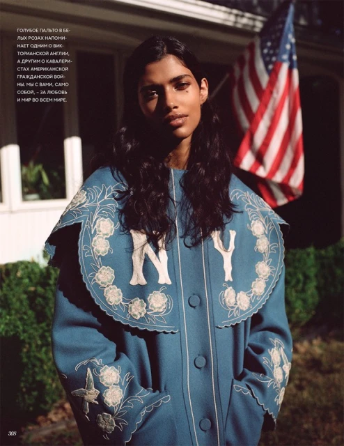 A young woman poses in a vintage-style blue coat embroidered with white flowers, standing outdoors with an American flag waving.