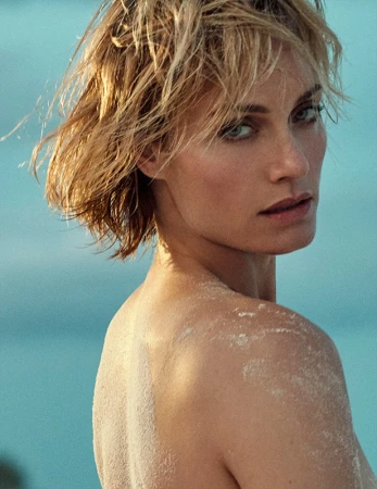 Close-up portrait of a woman with wet blonde hair looking over her shoulder, with sand on her bare shoulder.