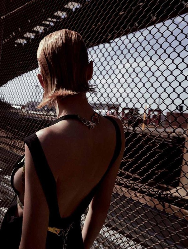 Back view of a woman with short hair wearing a black dress and necklace, standing behind a metal fence with an urban background.