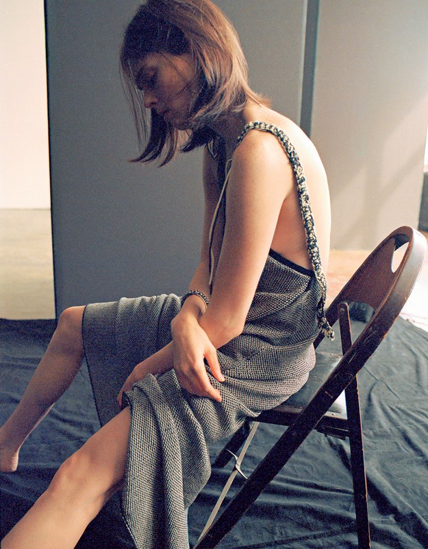 Young woman in gray dress sits on a folding chair indoors against a gray background, showcasing a fashion shoot with natural light.