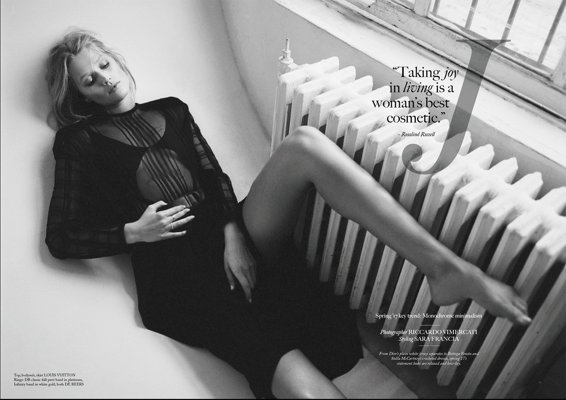 Black and white fashion shot of a blond adult female lying by a radiator in a sheer black dress, eyes closed, jewelry visible.