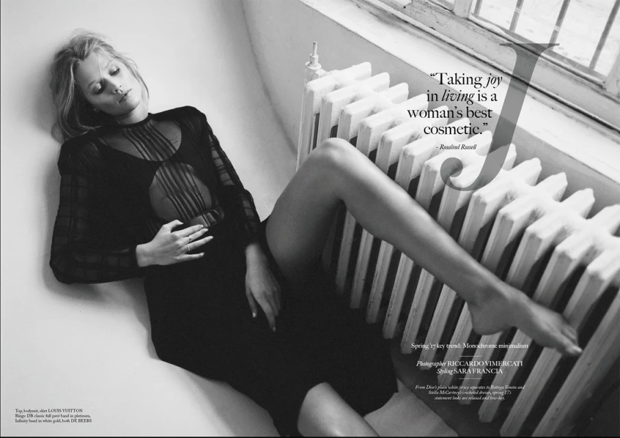 Black and white fashion shot of a blond adult female lying by a radiator in a sheer black dress, eyes closed, jewelry visible.