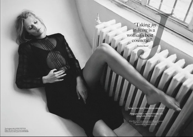 Black and white fashion shot of a blond adult female lying by a radiator in a sheer black dress, eyes closed, jewelry visible.