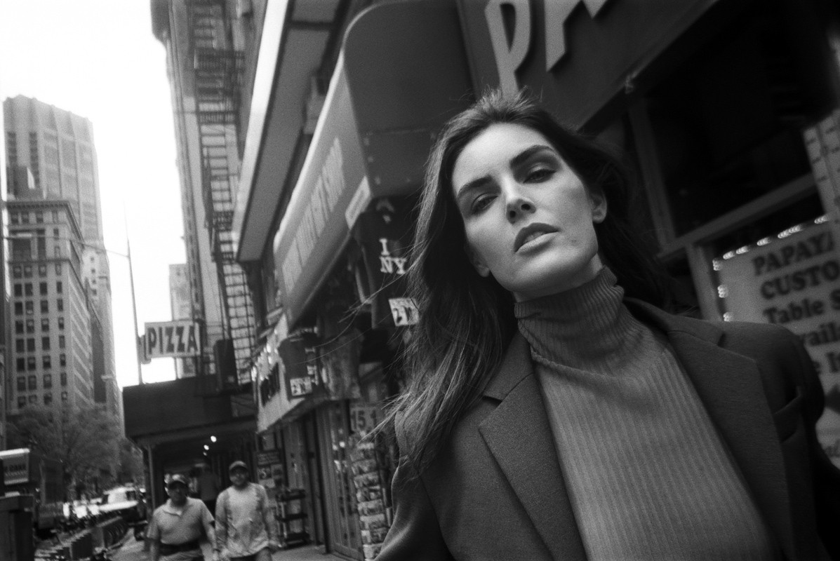 Black-and-white shot of a woman in a jacket on a city street with buildings, storefronts, and passersby visible in the background.
