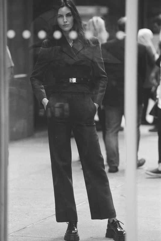 Monochrome image of a woman standing on a city street wearing a tailored suit and platform shoes, shot through a glass window.