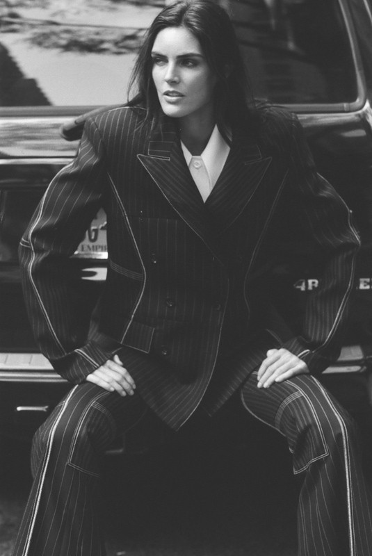 A brunette fashion model in a pinstripe suit sits atop the hood of a black car, posing with a serious expression in a black-and-white portrait.