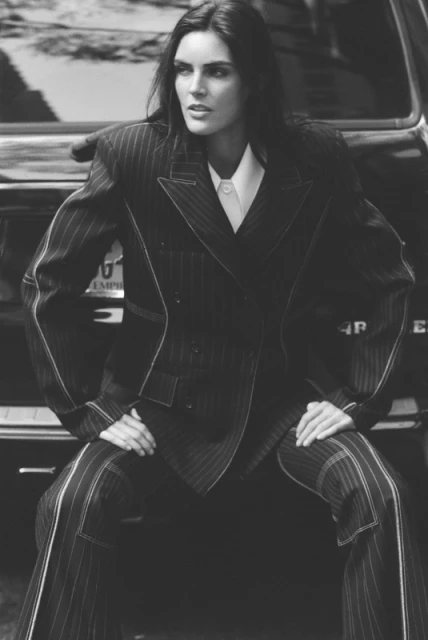 A brunette fashion model in a pinstripe suit sits atop the hood of a black car, posing with a serious expression in a black-and-white portrait.