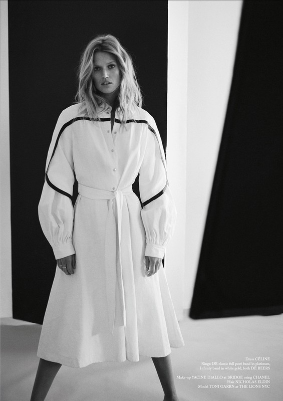 Black-and-white fashion shot of a blonde adult female in a white dress with black accents, posing against a graphic monochrome backdrop.