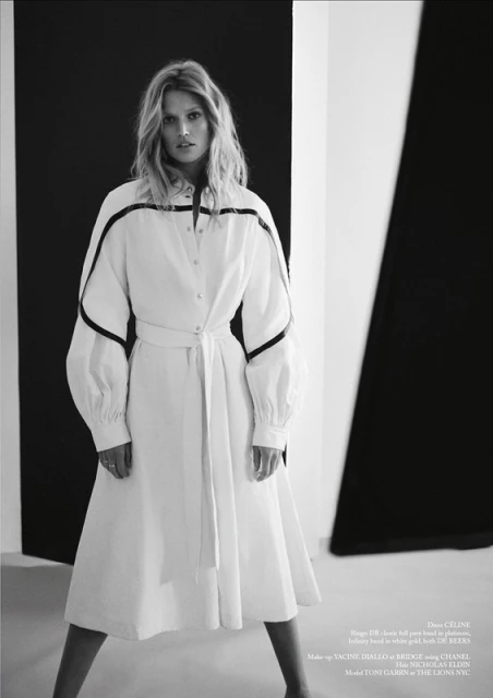 Black-and-white fashion shot of a blonde adult female in a white dress with black accents, posing against a graphic monochrome backdrop.