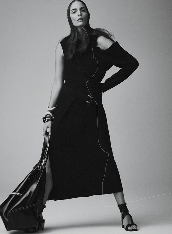 Monochrome fashion shot of a woman in a black dress and sandals, holding a black bag, posing against a neutral background.