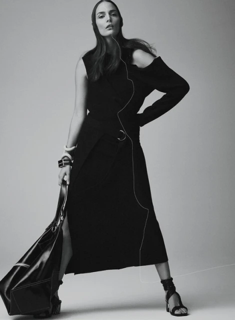 Monochrome fashion shot of a woman in a black dress and sandals, holding a black bag, posing against a neutral background.