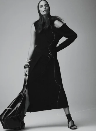 Monochrome fashion shot of a woman in a black dress and sandals, holding a black bag, posing against a neutral background.