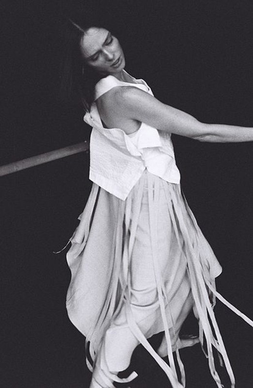 Woman in flowing white dress and top posing against a dark background, with a horizontal pole, shot in black and white.