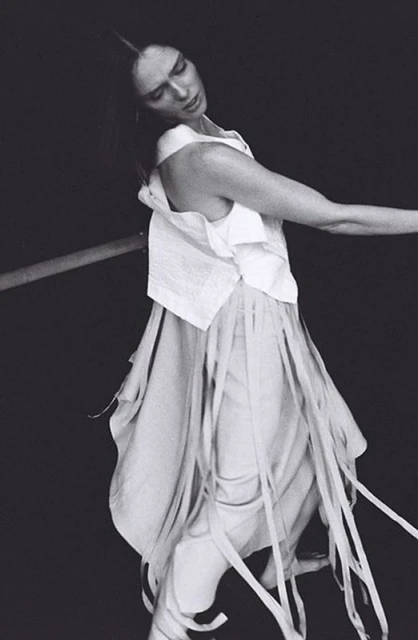Woman in flowing white dress and top posing against a dark background, with a horizontal pole, shot in black and white.