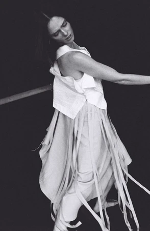 Woman in flowing white dress and top posing against a dark background, with a horizontal pole, shot in black and white.