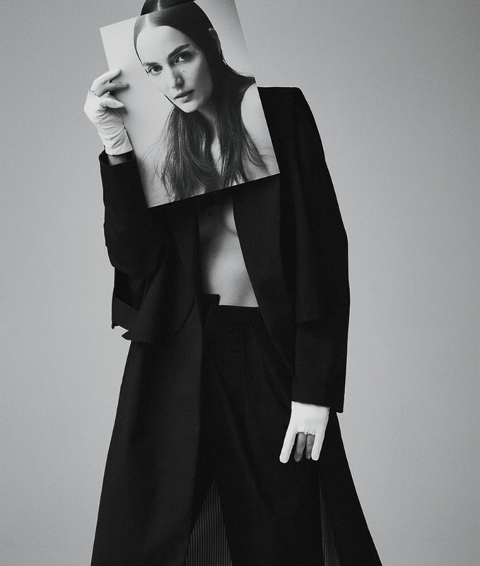 Woman in oversized black coat holds black and white portrait in front of her face in a studio setting with neutral background.