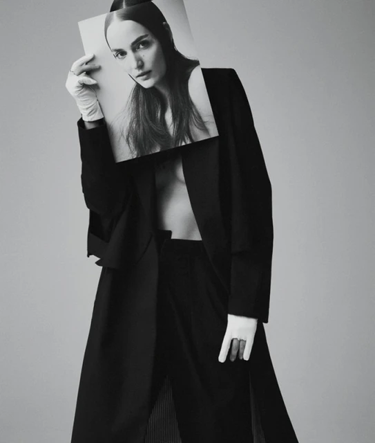 Woman in oversized black coat holds black and white portrait in front of her face in a studio setting with neutral background.
