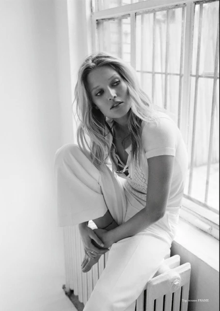 Black and white fashion shot of a blonde woman with long hair sitting on a radiator by a window in an indoor setting. 