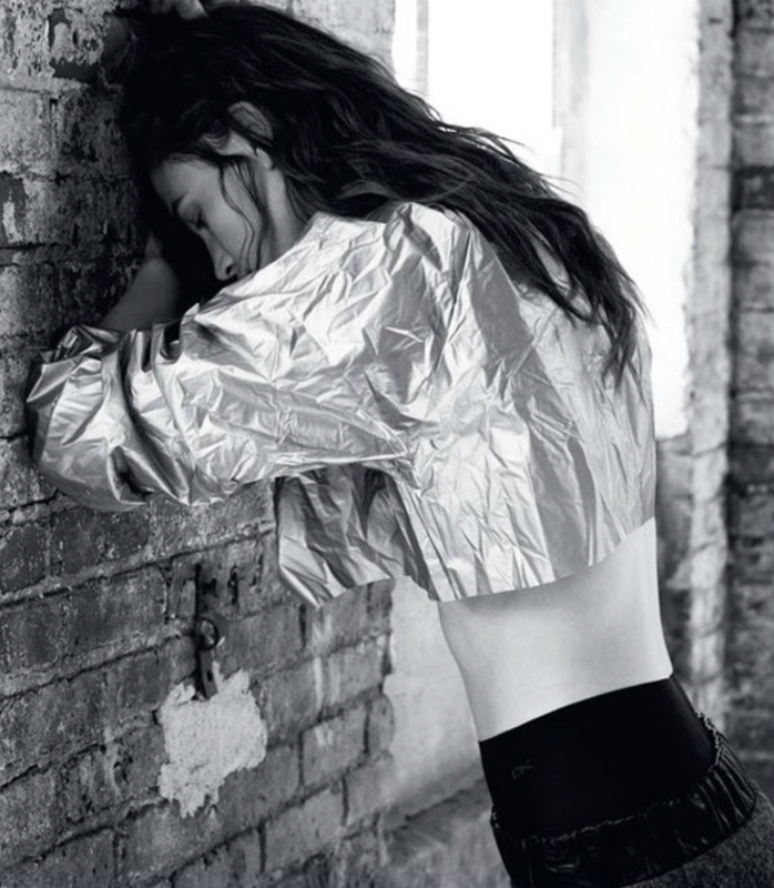A young adult woman with long brown hair, wearing a silver crop top, leans against a textured brick wall in a black and white studio shot.