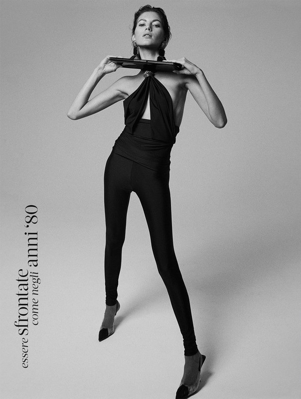 Fashion editorial shot in black and white of a young woman posing with dark hair and a dark top and leggings against a grey background.