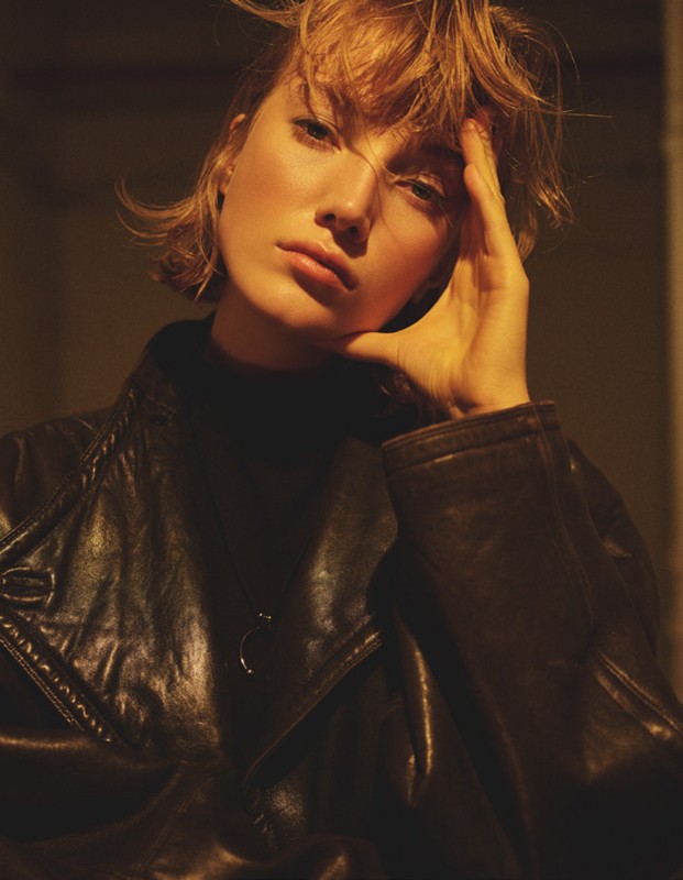Portrait of a young woman with short blonde hair wearing a leather jacket, posing indoors with a hand to her face in warm, subdued lighting.