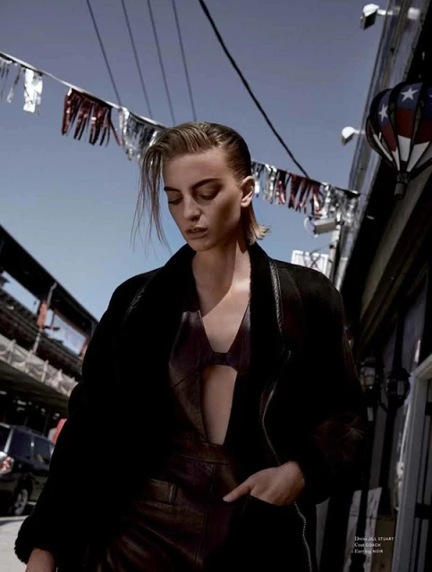 An adult woman with short hair poses outdoors in a black coat and leather overall dress, under American flag bunting and a blue sky.