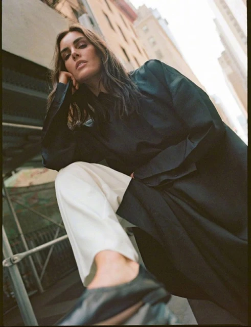 Woman poses on railing in black coat and white pants, with city buildings visible in the background, shot from low angle.