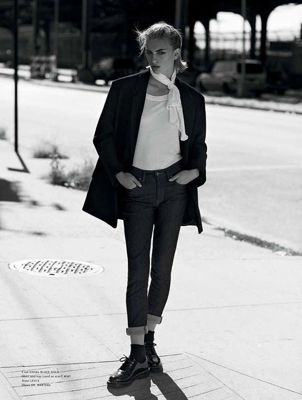 Black and white full-length portrait of a female fashion model on city street, wearing a coat, jeans, t-shirt, scarf, and platform shoes.