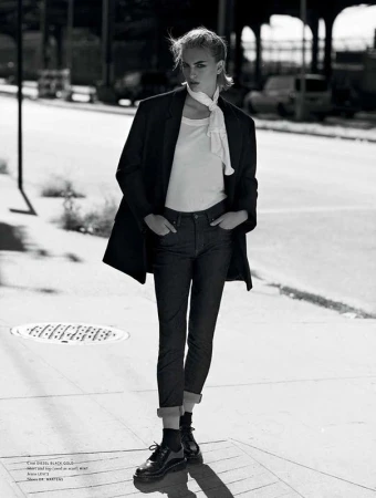 Black and white full-length portrait of a female fashion model on city street, wearing a coat, jeans, t-shirt, scarf, and platform shoes.