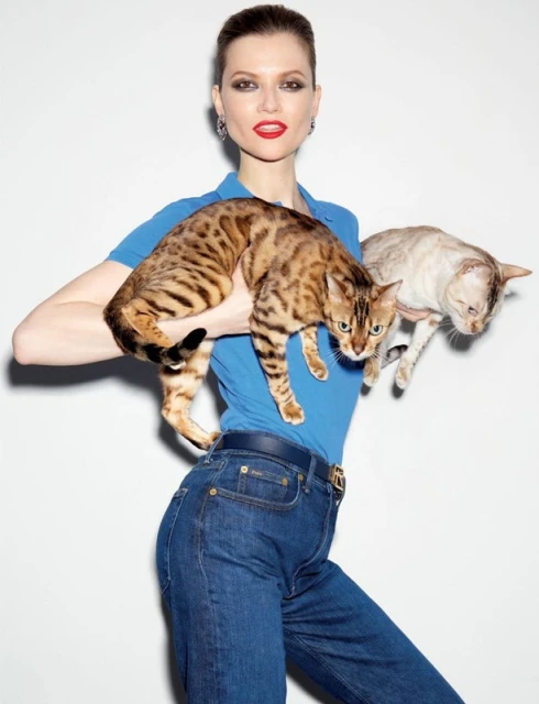 Adult woman holds two cats in a studio setting with a plain white background, wearing blue top and jeans. Cats are bengal and tabby.