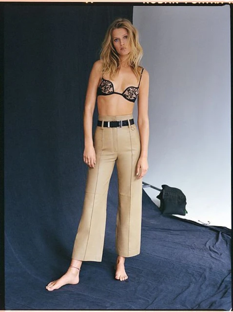 Blonde model poses in black lace bralette and khaki pants in a studio with dark blue and grey background, barefoot on blue material.
