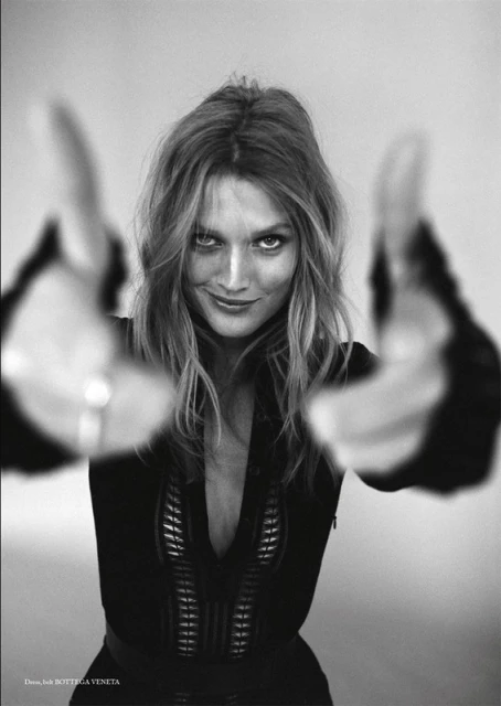 Black and white portrait of a woman with long wavy hair, smiling and holding her hands up to the camera in a photo studio.