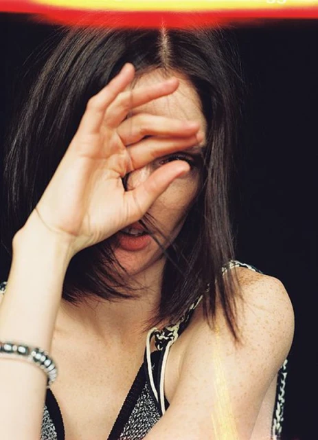 Close-up portrait of a woman with short brown hair partially covering her face with her hand against a dark background.