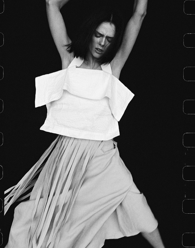 Black and white portrait of a woman in a white top and light skirt with dark hair dancing with her arms above her head.