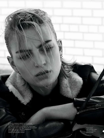 Close-up monochrome portrait of a model with slicked-back hair wearing a leather jacket with shearling collar against a light brick wall.