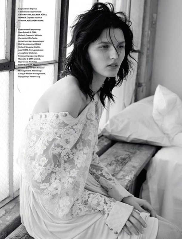 Black and white editorial portrait of a woman with tousled dark hair wearing a lace dress near a window