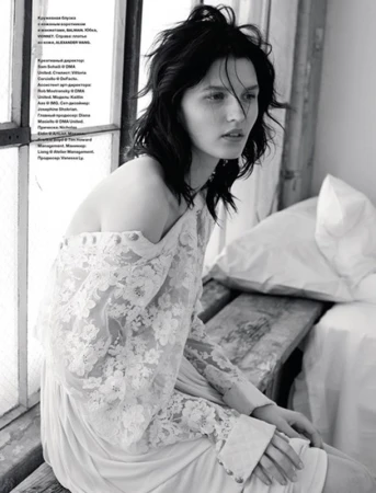 Black and white editorial portrait of a woman with tousled dark hair wearing a lace dress near a window