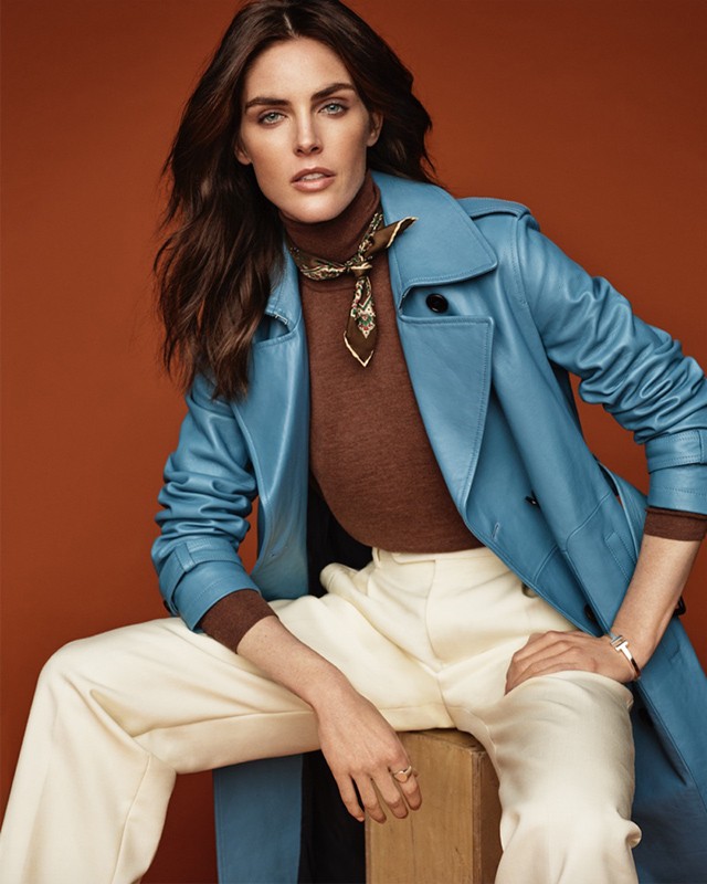 A brunette woman in a blue coat poses against a burnt-orange backdrop, seated on a wooden block wearing a scarf and white trousers.