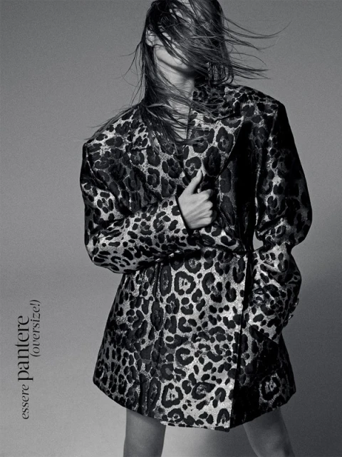 Model wearing an oversized leopard print blazer with hair blowing across face against a grey background.