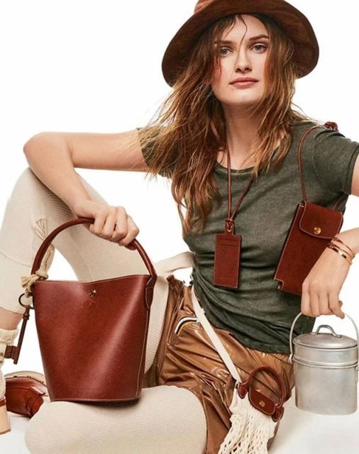 Woman in a brown hat and green t-shirt holding a brown leather bucket bag against a plain white background