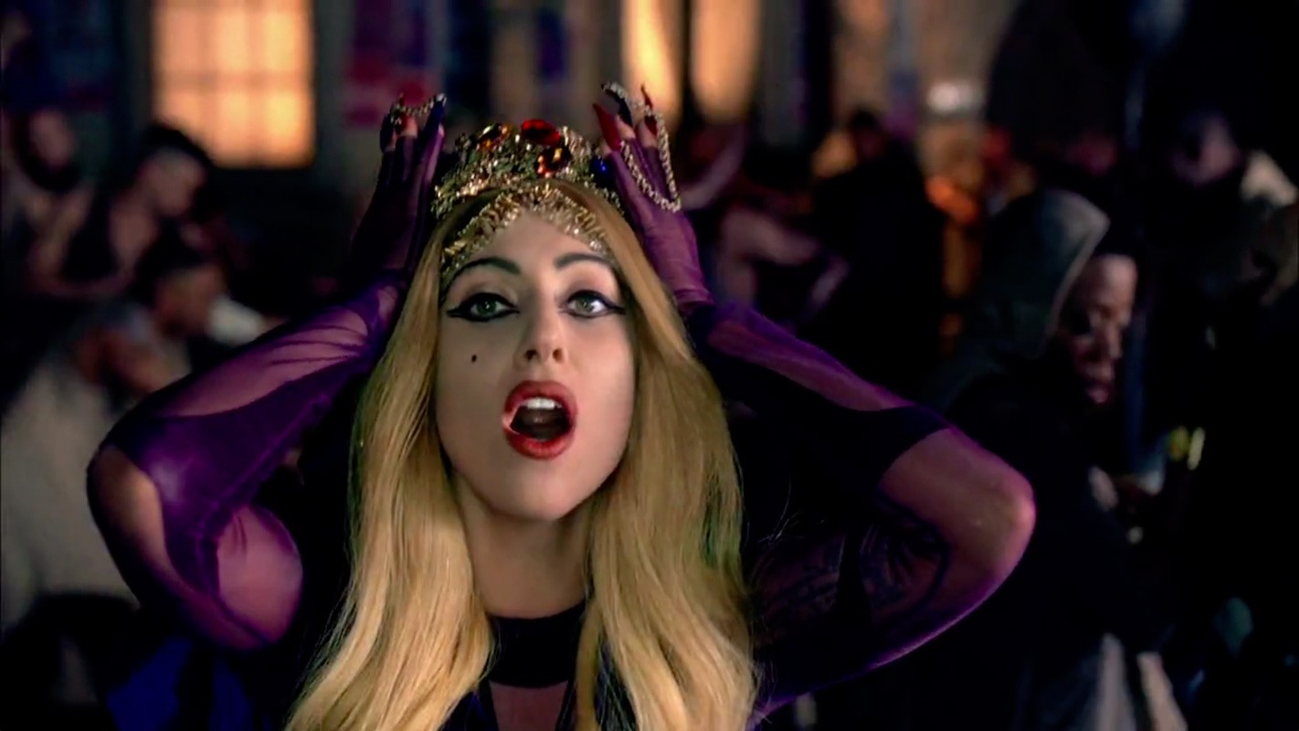 Close up of Lady Gaga wearing a crown and purple gloves surrounded by a crowd of people in a dimly lit indoor setting.