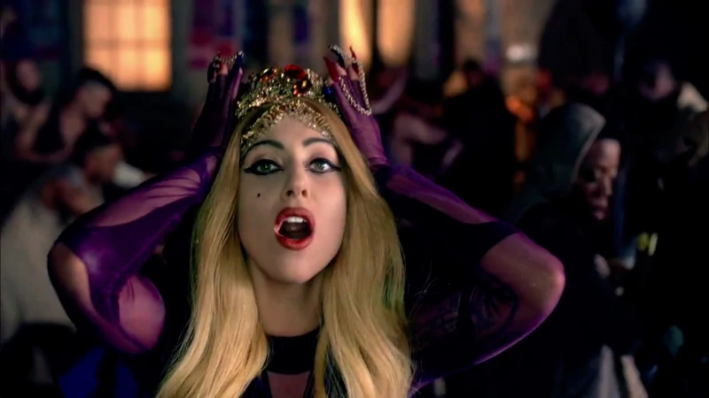 Close up of Lady Gaga wearing a crown and purple gloves surrounded by a crowd of people in a dimly lit indoor setting.