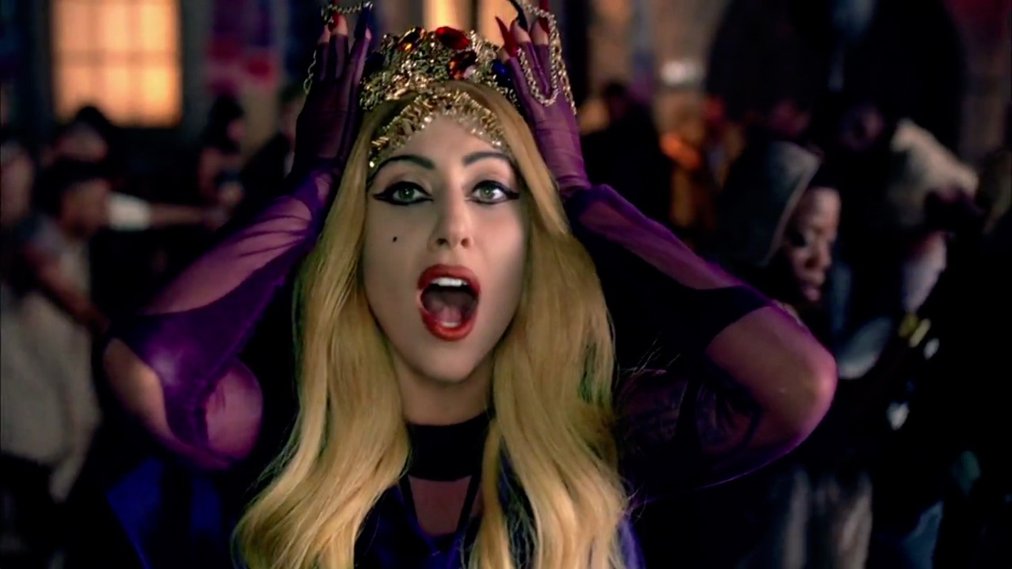 Close-up of an adult female vocalist with open mouth and a crown, wearing purple gloves in front of a dark, blurred crowd.