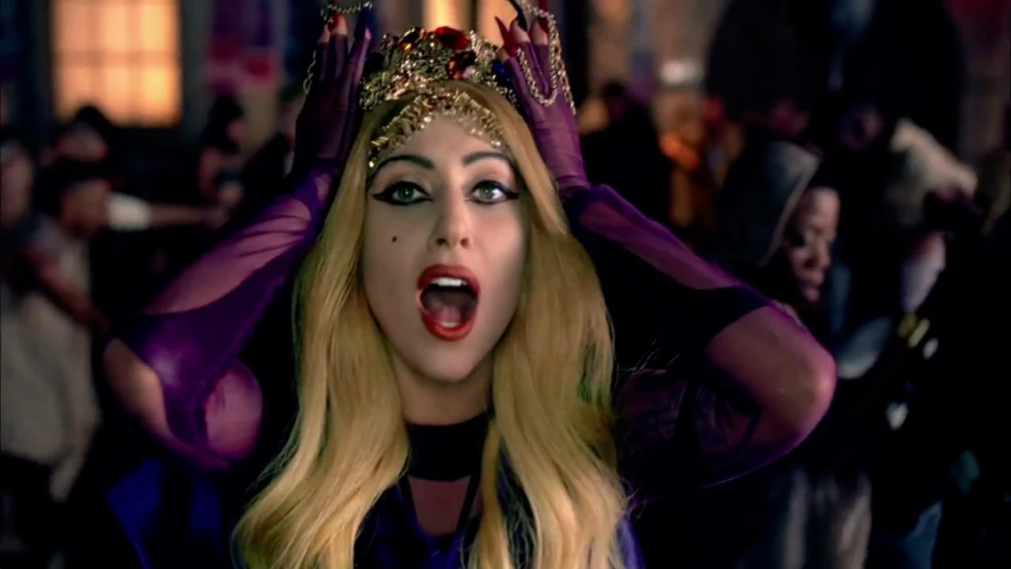 Close-up of an adult female vocalist with open mouth and a crown, wearing purple gloves in front of a dark, blurred crowd.